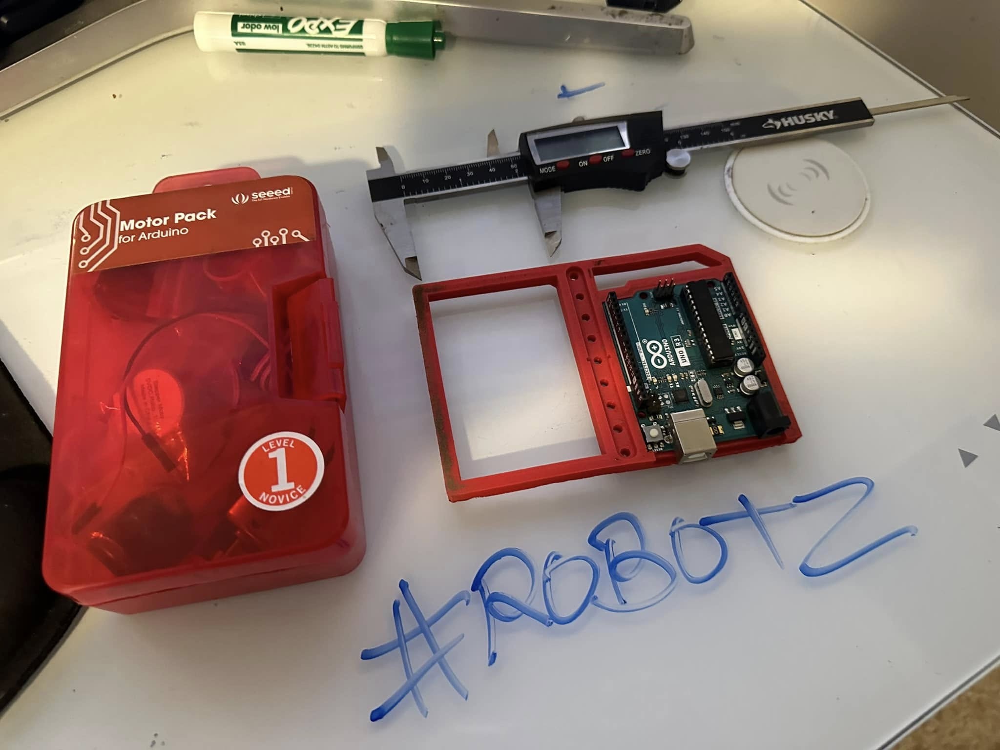 Arduino Uno fitted into a measured red enclosure beside digital calipers, motor components, and working notes